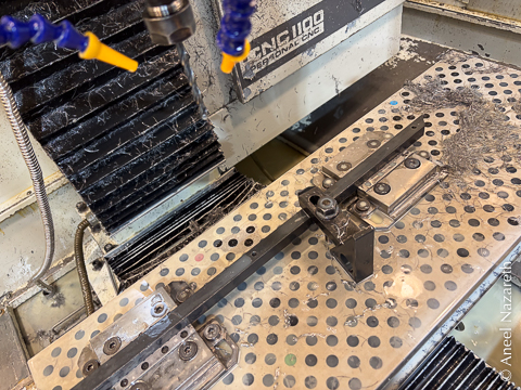 A bar in a CNC milling machine. The bar is clamped to the bed using two vises and held down by a strap clamp